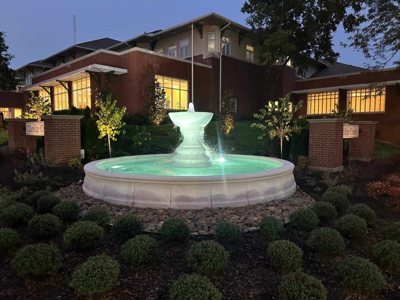 Illuminated stone fountain at Claridge Court Retirement Community in Prairie Village, Kansas, surrounded by landscaped garden at dusk