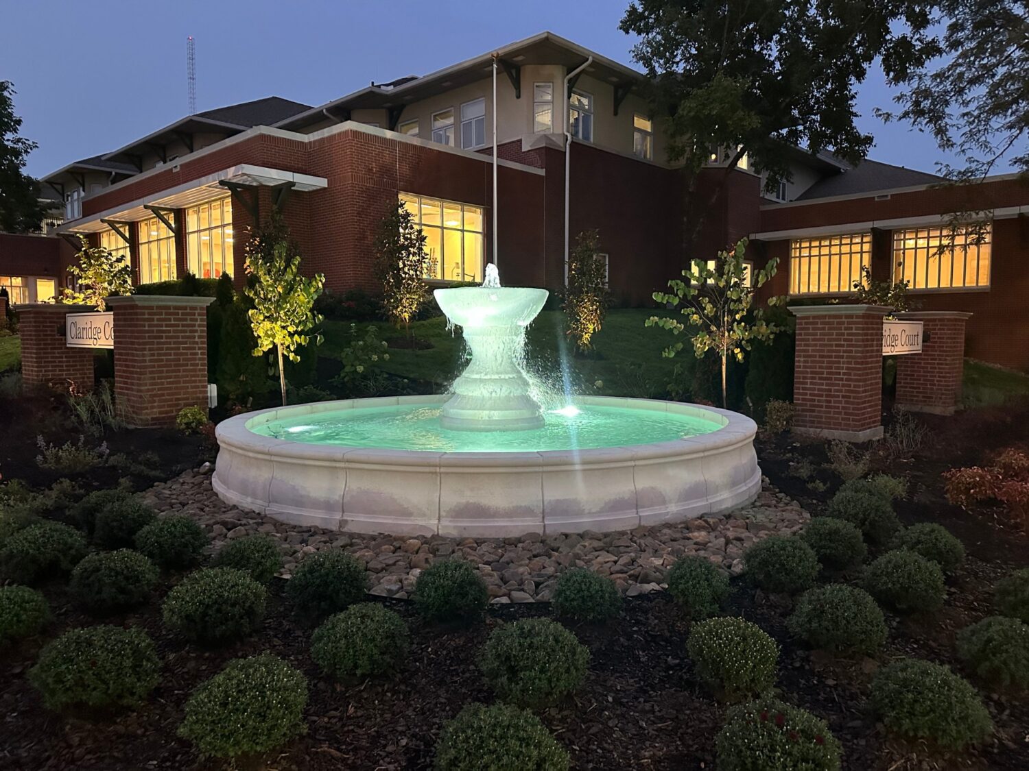 Illuminated stone fountain at Claridge Court Retirement Community in Prairie Village, Kansas, surrounded by landscaped garden at dusk